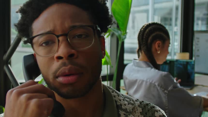 In an office a man talks to his coworker and then goes back to talking on the phone with a confused facial expression