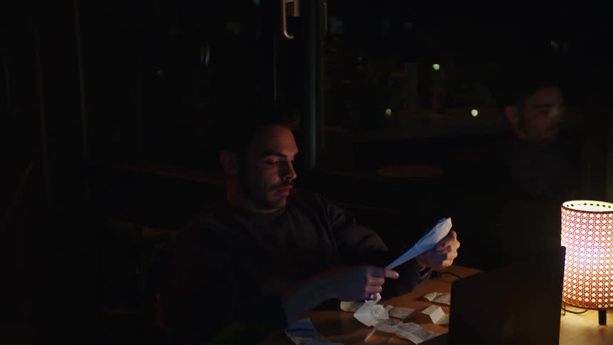 Man with tired facial expression receiving an invoice receipt from a woman while working on his laptop at night