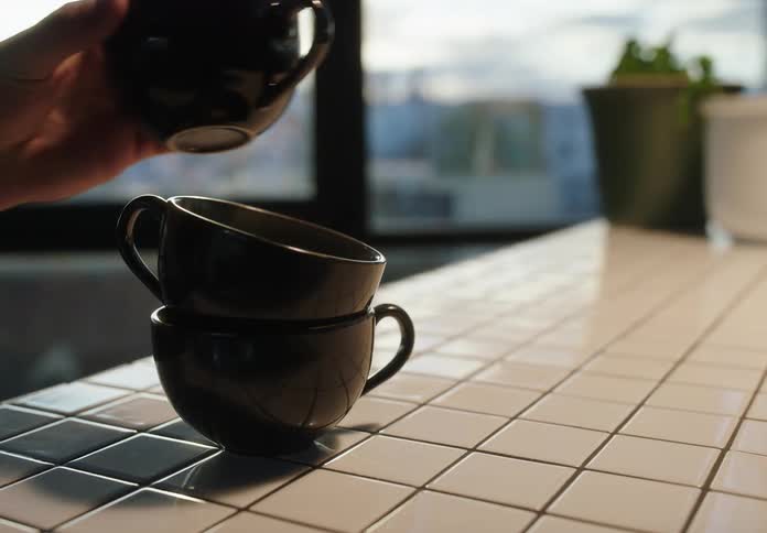 Four brown coffee cups being stacked together one at a time on top of a white tile counter top with a large window in the background
