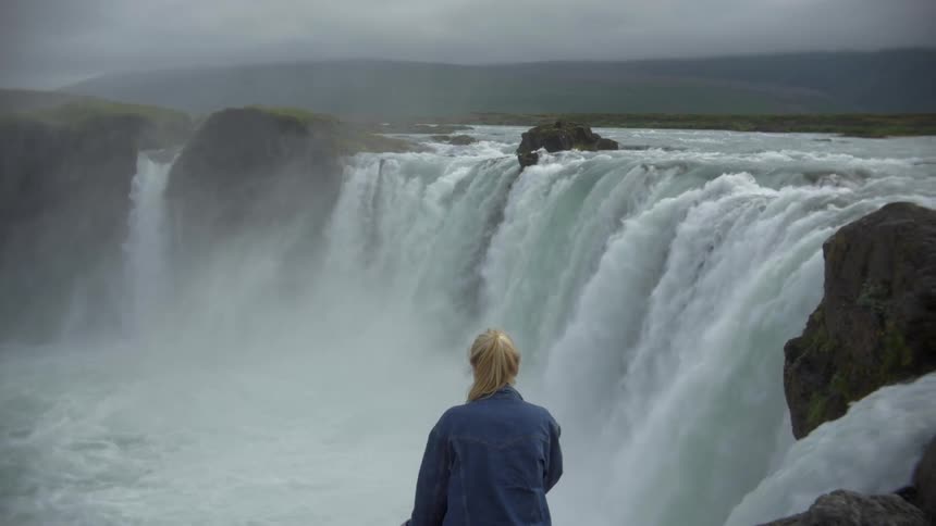 A woman is standing by a waterfall and looking at it