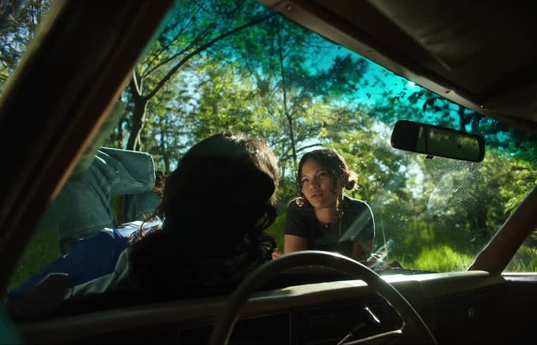 Two women on the hood of a car with one of them lying down smoking