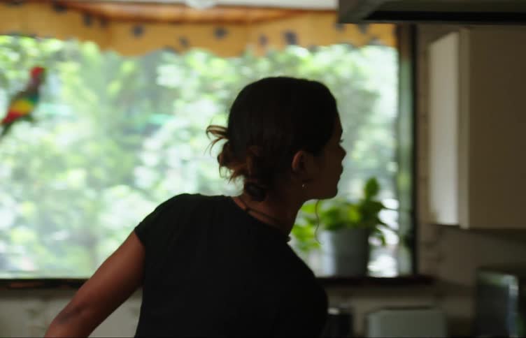 A young woman is standing in the kitchen when she hears something outside that catches her attention and leaves to check it