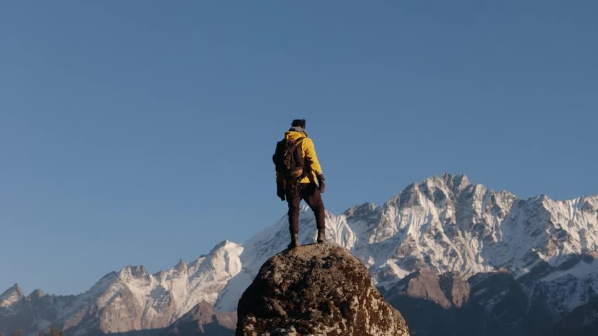 Backpacker stands on rock outcropping and looks at snowy mountains