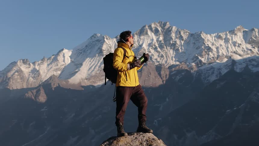 Man on a rock peak on a cliff taking a sip of water while having an idyllic view of mountains with snow all around him
