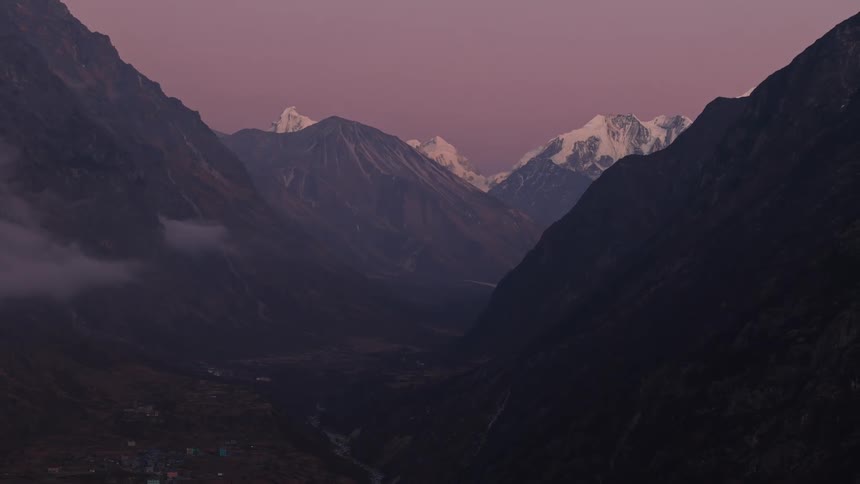 An aerial view of a valley between large mountains and in the background there are mountains with their tops covered in snow