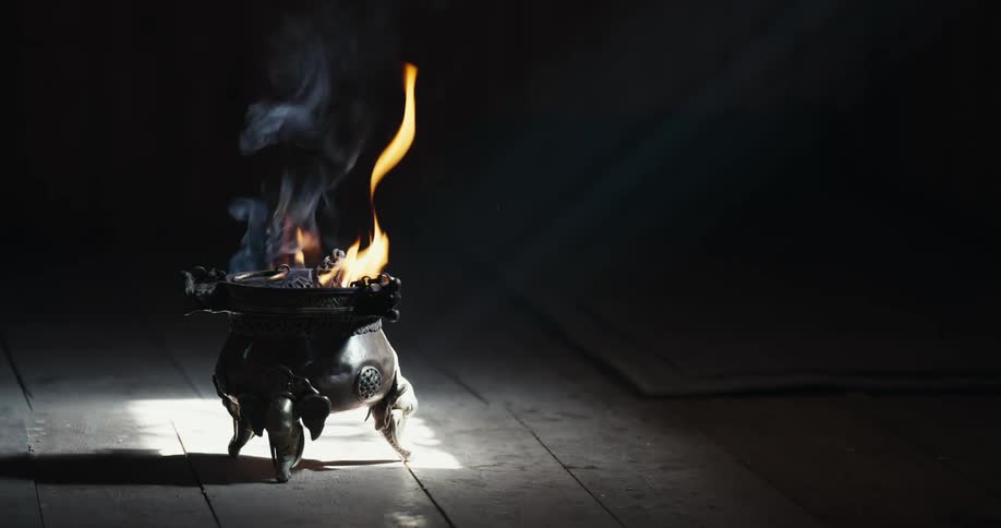 A cauldron is on the wooden floor and is receiving sunlight while it is smoking and burning something on top of it