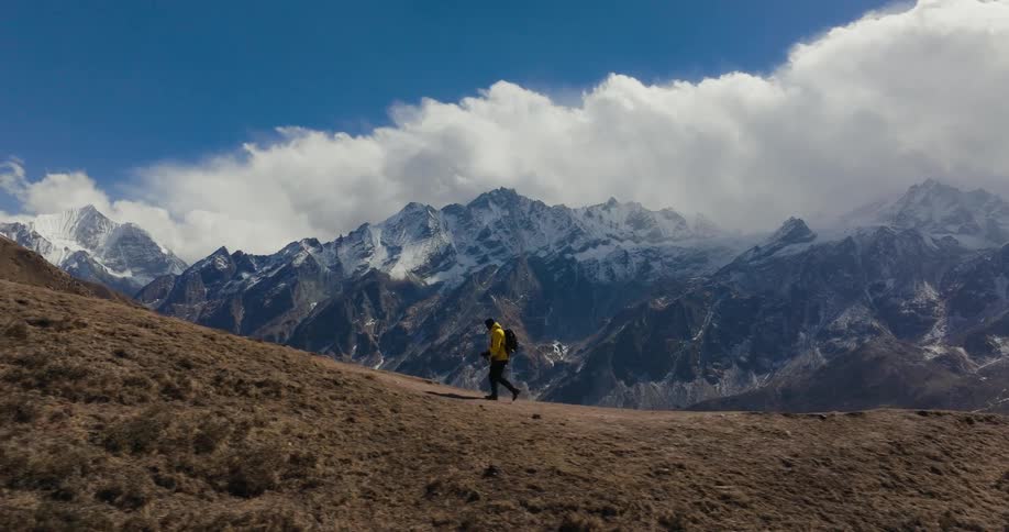 A person in a yellow jacket is walking on top of a mountain in a montagnesque region with huge white clouds in the sky