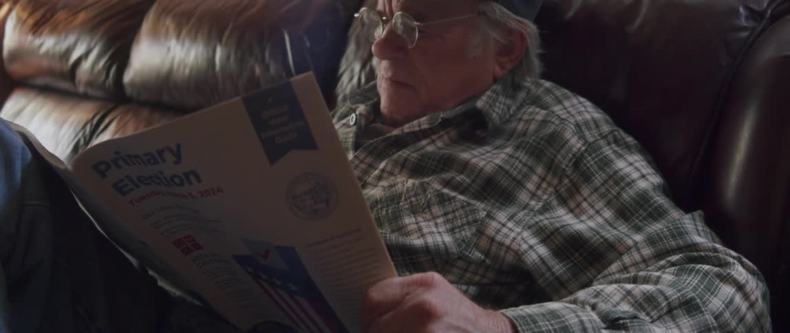 An old man is sitting on a sofa while reading a magazine and he is wearing reading glasses and a cap