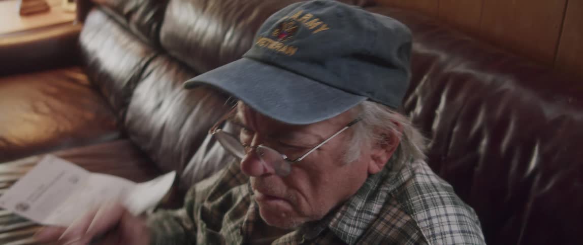 An elderly man is sitting on the sofa while reading a magazine and then takes off his glasses and puts them back on