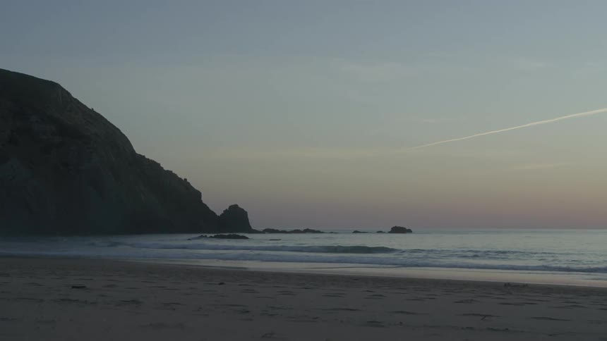 A beach with a mountain on the left side and small waves reaching the coast and the sky has pink tones