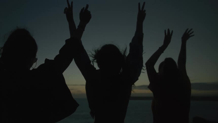 Silhouette of three women with their backs facing the sea with their hands raised in a dark dawn sky