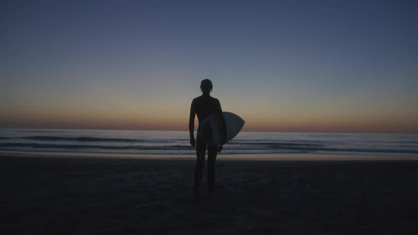 A man watches the sunrise while holding his surfboard