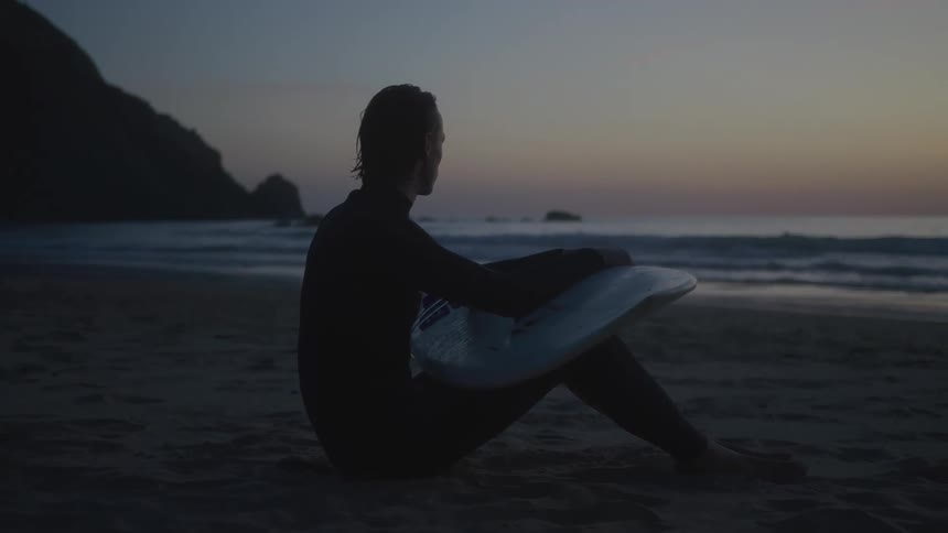 A surfer sits on a sandy beach while holding his surfboard and watching the ocean