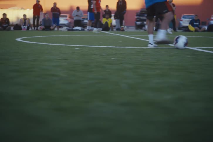 Teenagers playing football on a society field in an outdoor setting with people around watching