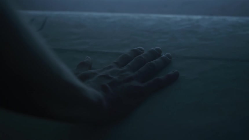 A hand slowly descends in the dark to touch a surfboard on a table