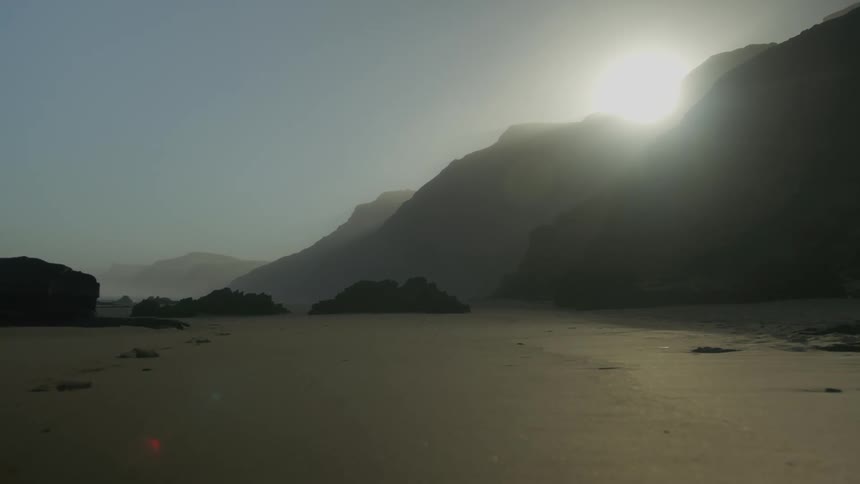 A beach scene with sunlight breaking over the mountain through fog