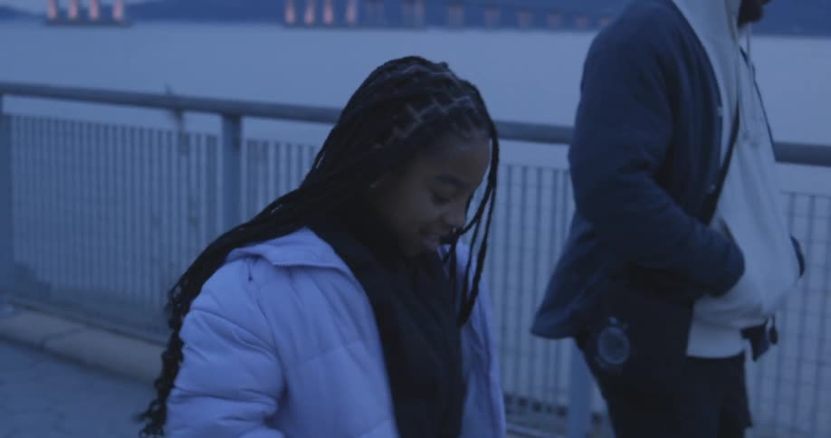 A girl next to a boy and they are both walking and close to a handrail facing the water