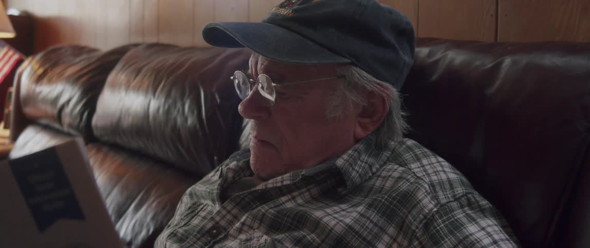 An elderly man in glasses and a cap is sitting while reading a magazine