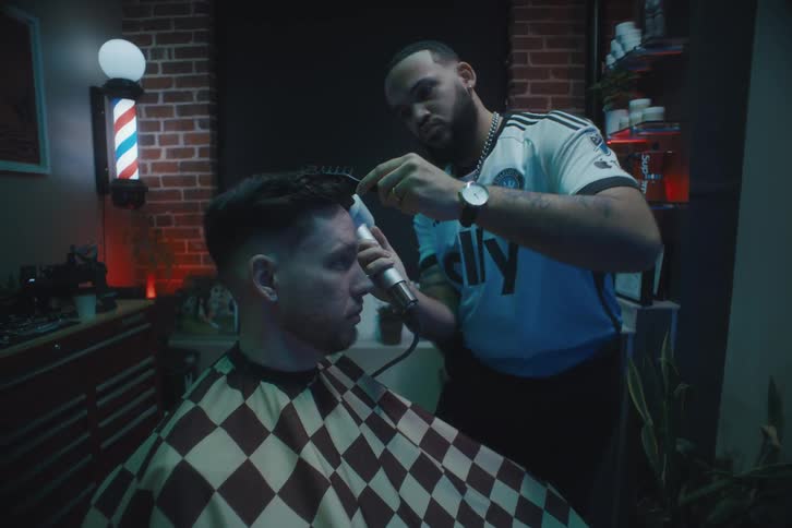 A barber is combing and cutting the hair of a man who is sitting wearing a checkered sheet