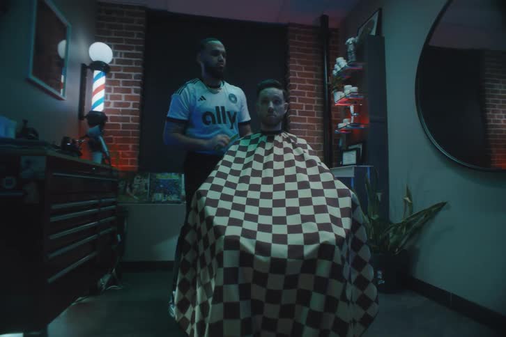 A barbershop where there is a man sitting with a sheet on his body and the barber turning the chair and combing the sitting mans hair