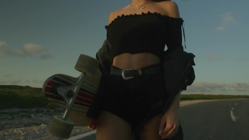 A scantily dressed woman walking along a road carrying a skateboard