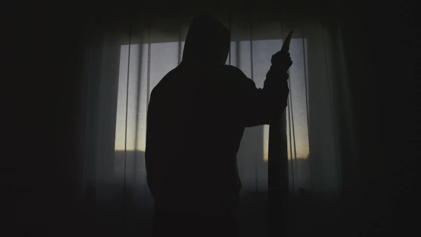 Man holding long pointed object stands silhouetted against curtained window