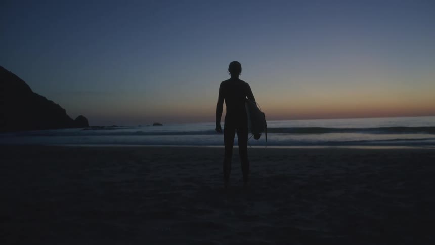 A surfer is standing on the beach with his surfboard watching the sunrise