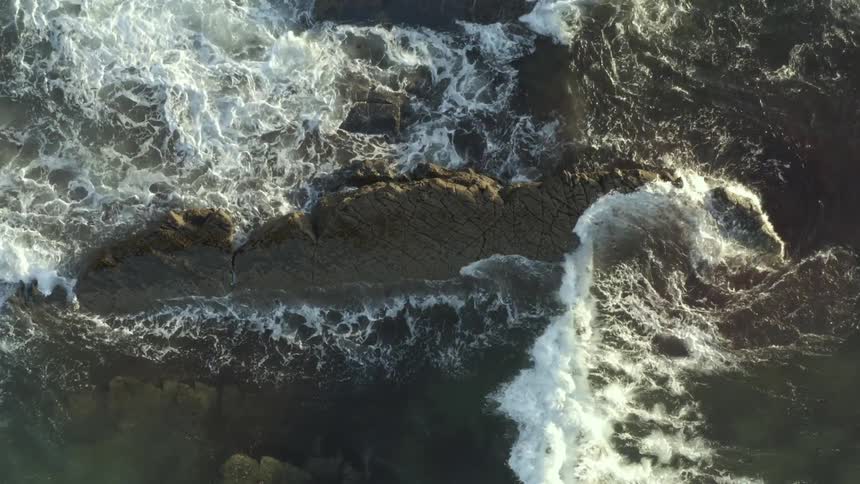 Numerous waves push past an elongated rocky prominence at the coast