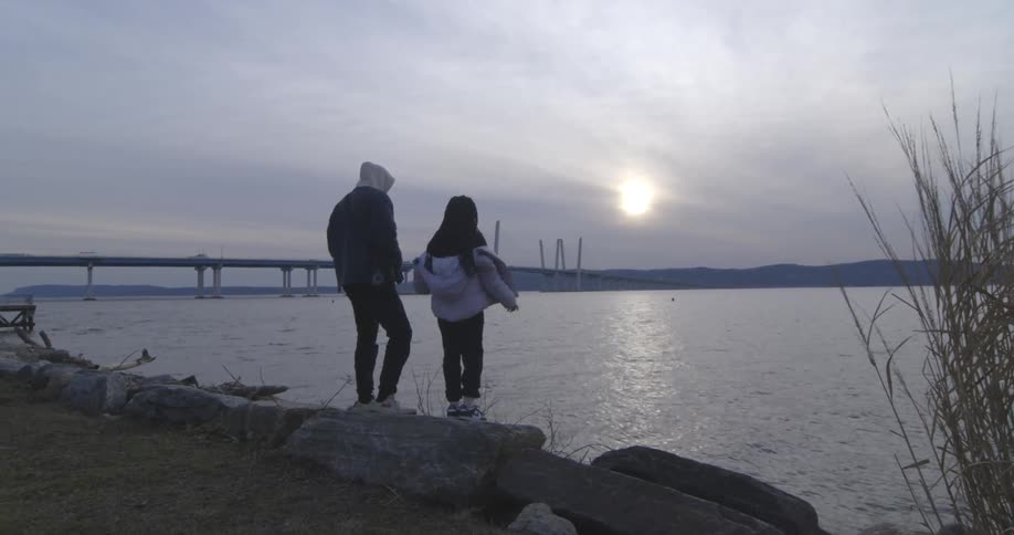 Father next to his little daughter are standing on a rock in front of the sea with a sun on the horizon