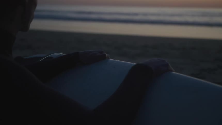 A person holding a surfboard is sitting on the sand watching the sea from the beach