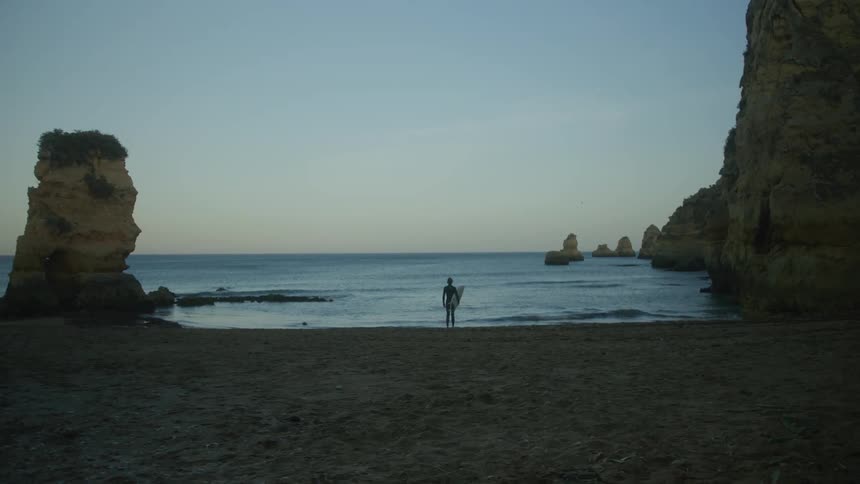 Man stands on a beach holding a surfboard watching waves roll in to shore