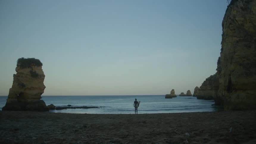 Landscape of a beach on a morning with rocks around and a person holding a surfboard on the shore