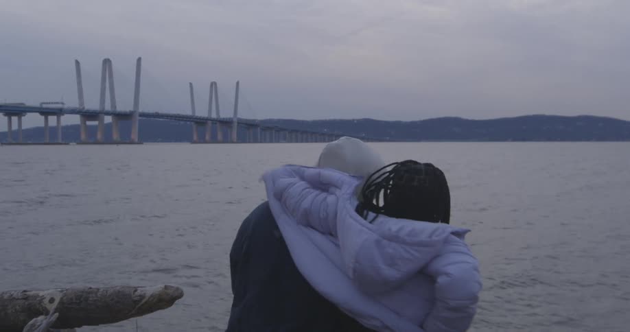 Two people sit by the shore near the bridge