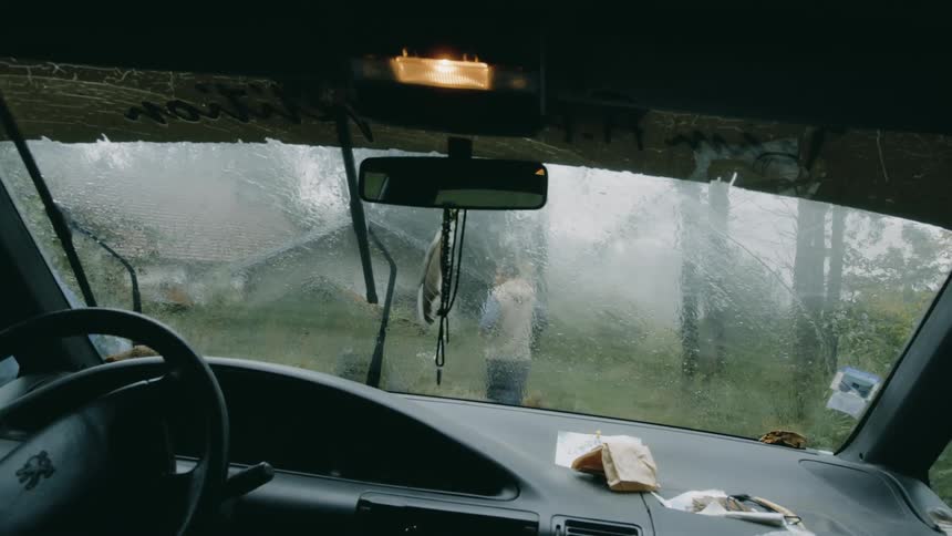 Interior view of a vehicles windshield with a person outside walking towards a house on the farm on a rainy day