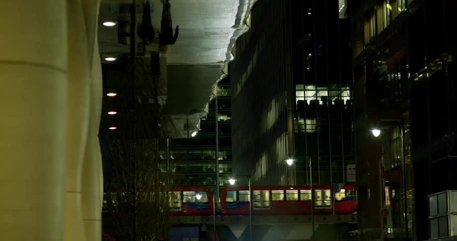 Train passing over a city at night with two people walking on a sidewalk and flaring car headlights