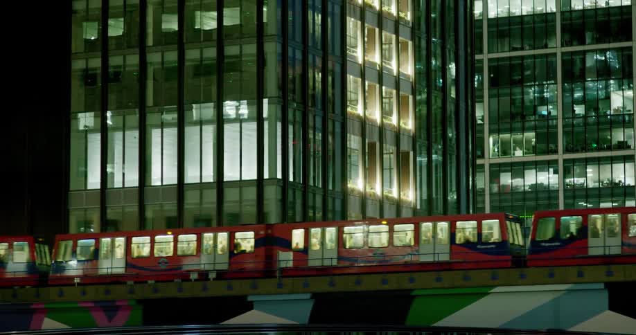 Red train passing on a viaduct next to modern buildings at night