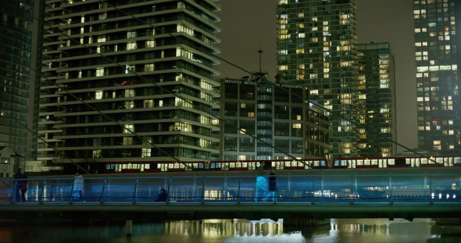 Several people walking along a walkway next to several buildings and there is a train passing behind