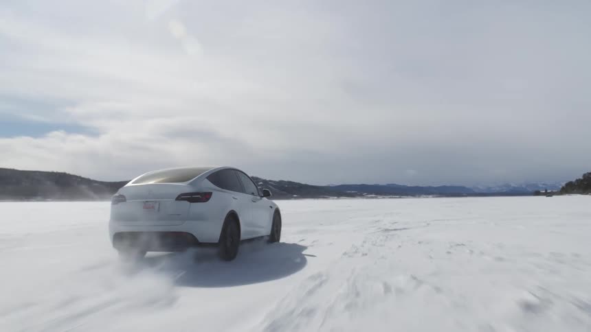 A white car drives over a frozen lake covered in snow while snow blows by