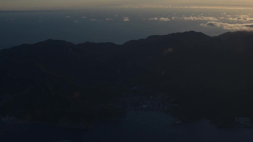 An aerial view of a huge island with a city facing the water and white clouds in the sky that are receiving sunlight
