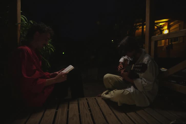 Two men are sitting on a wooden porch and one of them is playing the guitar and the other is reading a book