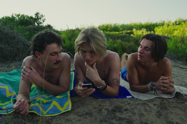 Three young men lie on the ground while one uses a smartphone