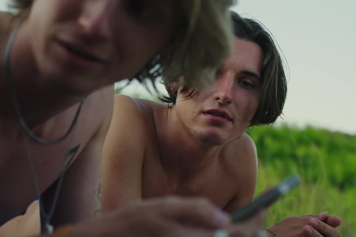 Two boys are looking at a cell phone and one of them is showing the screen to someone else and talking to them