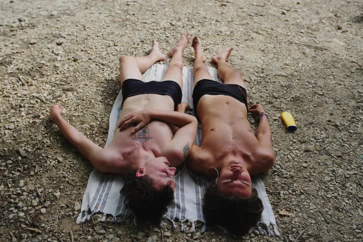 Two men in just swim trunks are lying on a towel next to each other while listening to music on headphones and talking and then one gets up and leaves