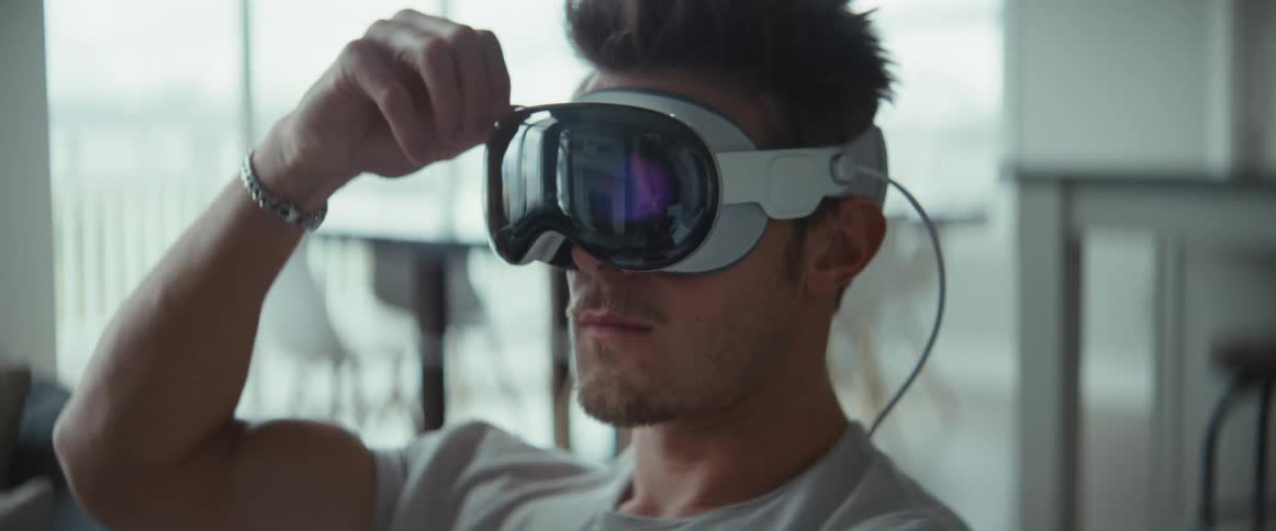 A man is sitting wearing a virtual reality glasses on his face and is adjusting it with his finger