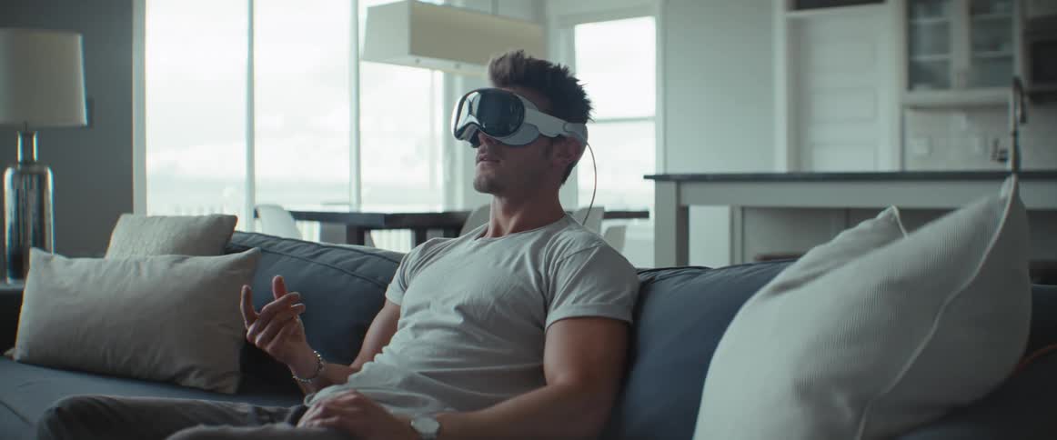 A man is sitting on the sofa while wearing a virtual reality glasses on his face and moving his fingers in a claw shape