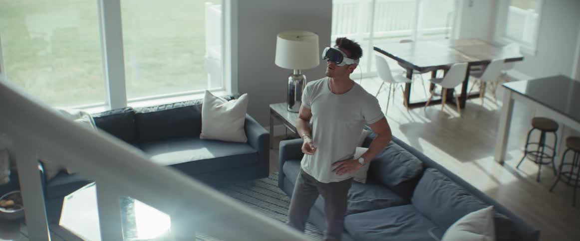 A man is standing in a living room with a blue sofa and he is talking and smiling while wearing a virtual reality goggle on his head