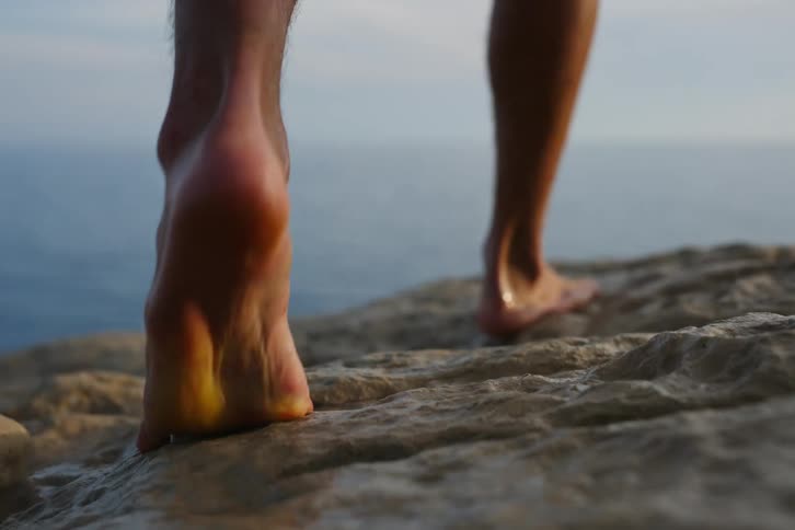 A mans feet are on a rocky surface and then he starts walking and jumps towards the water