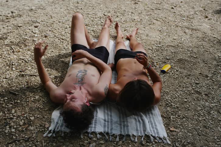 Two friends in swim trunks lying on a towel sharing the same headset and relaxing