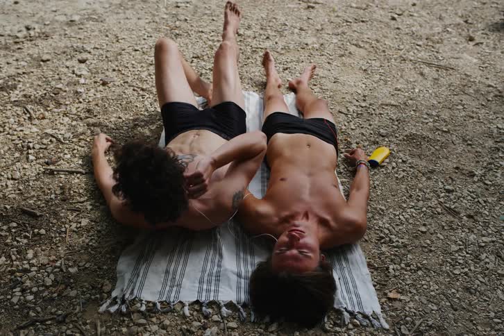 Two men sharing headphones while sunbathing talk then one of them gets up and walks away