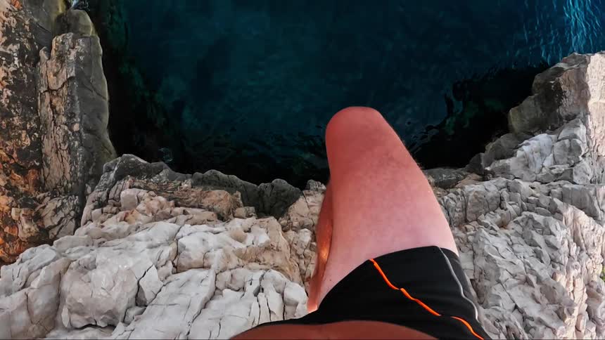 Young man stands on the edge of a rocky cliff then jumps falling into the water in slow motion